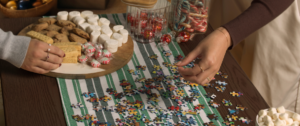 Two hands working on a colorful jigsaw puzzle spread across a green-striped cloth, surrounded by festive treats including shortbread, chocolate chip cookies, peppermint candies, marshmallows, and jars filled with candy canes and Lindt truffles.