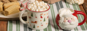 Two festive mugs on a striped green and white cloth: one white mug with holiday patterns filled with mini marshmallows, and one Santa-shaped mug topped with whipped cream, surrounded by shortbread cookies and peppermint candies.