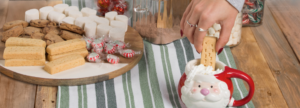 Hand dipping a shortbread cookie into a Santa-shaped mug filled with whipped cream, next to a wooden tray with assorted holiday treats including chocolate chip cookies, marshmallows, and peppermint candies on a green and white striped cloth.