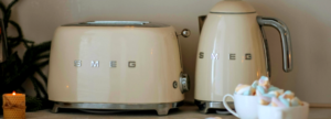 Cream-colored SMEG toaster and electric kettle on a countertop with festive greenery, a lit candle, and a white cup filled with pastel marshmallows in the foreground.