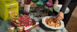 Festive holiday table with a green tree-patterned tablecloth featuring a box of Panettone, a red gift box with gold ribbon, two gold-wrapped chocolate bears, a bottle of rosé wine, colorful stemmed glasses, and a decorative bowl of assorted chips with a hand reaching in.