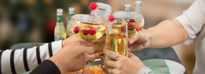 Four hands clinking glasses of sparkling mocktails garnished with cranberries and raspberries, with bottled drinks in the background on a festive table setting.