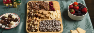 Festive snack board with assorted chocolates, fudge, crackers, nuts, and pretzels, alongside a plate of truffles and a bowl of fresh berries on a holiday-themed tablecloth.