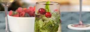 Close-up of a festive mocktail in a clear glass, garnished with fresh mint leaves and bright red cranberries, with a blurred background showing a bowl of mixed berries and a wine glass.