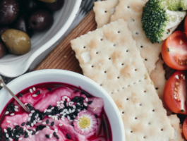 A vibrant vegetarian snack spread featuring a dip with sesame seeds and edible flower, fresh avocado slices, cherry tomatoes, broccoli florets, carrot sticks, saltine crackers, and a bowl of mixed olives on a rustic wooden surface.
