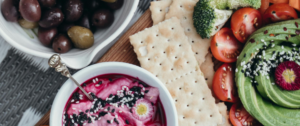 A vibrant vegetarian snack spread featuring a dip with sesame seeds and edible flower, fresh avocado slices, cherry tomatoes, broccoli florets, carrot sticks, saltine crackers, and a bowl of mixed olives on a rustic wooden surface.