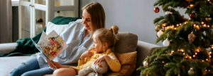 Person sitting on a sofa reading a colorful children’s book to a child holding a stuffed toy, next to a decorated Christmas tree with glowing lights and ornaments.