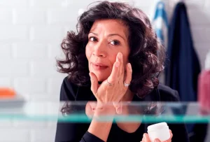 A person with curly dark hair applies skincare cream while looking into a bathroom mirror, holding a small white jar in one hand.