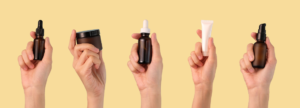 Five hands holding various skincare containers, including dropper bottles, a jar, and a pump tube, against a yellow background.