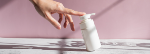 Hand reaching toward a white pump‑top skincare bottle on a light surface with a pink background.