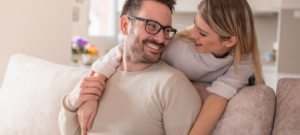 Two people sitting on a couch in a home environment, with one person placing an arm on the other’s shoulder to offer support.